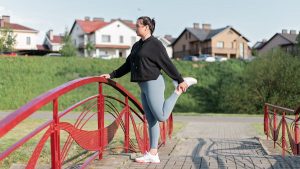 A woman in active wear performs a stretching exercise on a red bridge in a suburban park setting.