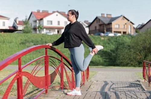 A woman in active wear performs a stretching exercise on a red bridge in a suburban park setting.